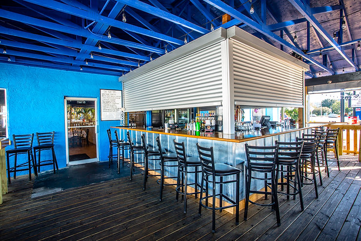 661c5d_23ee9d34ba624cf58640ca525 Modern outdoor bar in Morehead City with high chairs, blue ceiling beams, and metallic accents on a wooden floor—designed with Roll Shutters for added storm protection.