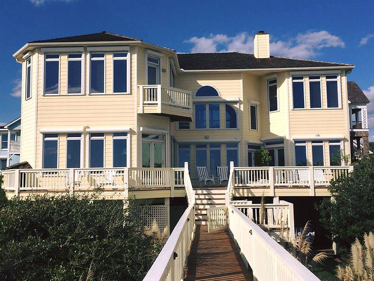 661c5d_75a78908c18a4b548b4e6acf3 Large yellow beach house with many windows, storm shutters for added protection, a deck, and a wooden walkway leading to the entrance—perfect for coastal living in North Carolina.