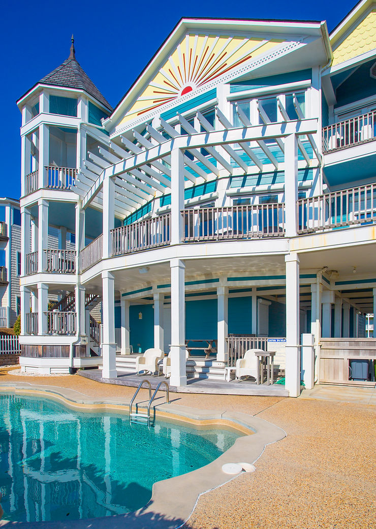 Large, colorful Atlantic Breeze beach house with balconies and a pool in front under a bright blue sky, featuring Roll Shutters for storm protection.