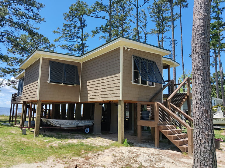 661c5d_7dc3778518ea4cacbe72aa01b Elevated beach house with wooden siding, large windows, and storm shutters, a boat stored underneath, surrounded by tall trees—perfect for catching the Atlantic breeze in North Carolina.