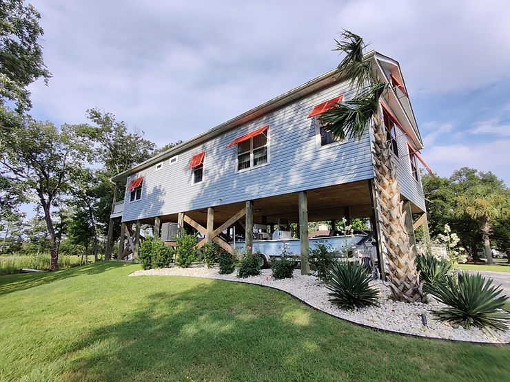 661c5d_80282840d97149bcb4789acbe Blue elevated house on stilts with red storm shutters for added storm protection, palm trees, and green lawn under a partly cloudy North Carolina sky.