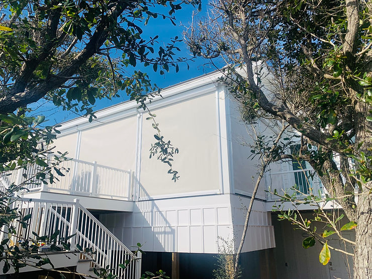 White house with a balcony and staircase, partially obscured by trees under a clear blue sky in Morehead City, features Roll Shutters for added storm protection.