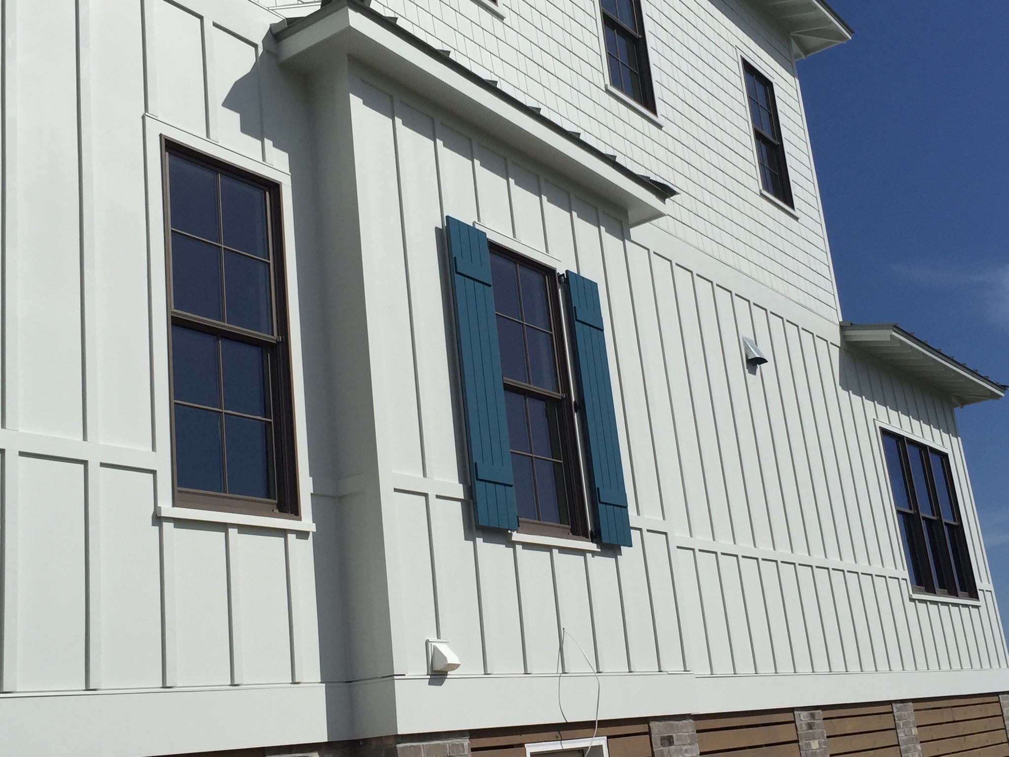 ATLANTIC BREEZE STORM SHUTTERS COLONIAL SHUTTERS1 White house with vertical siding, two windows, one with blue shutters, under a clear blue sky.