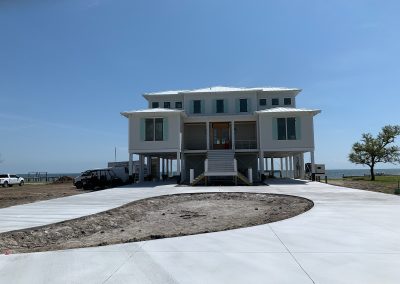 White elevated beach house on stilts with a circular driveway, near the ocean under a clear blue sky in Morehead City, North Carolina. Equipped with roll shutters for added protection.
