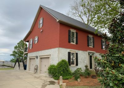 Red house with black shutters and garage doors, surrounded by lush greenery under a cloudy sky. Outfitted with Roll Shutters for added storm protection, this home offers comfort and safety against the Atlantic Breeze.