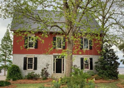 A two-story red and white house with black shutters provides storm protection and is partly shaded by a leafy tree in front, nestled in North Carolina.
