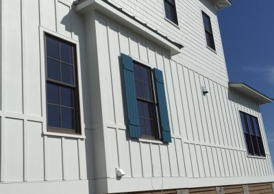 White house exterior with vertical siding, blue storm shutters, and black framed windows against a clear Atlantic Breeze under a clear blue sky.
