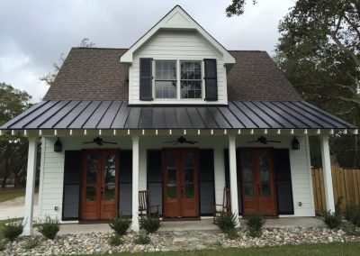 A white two-story house with dark shutters, a metal roof, and three wooden front doors with rocking chairs features Roll Shutters for added storm protection, perfect for North Carolina weather.