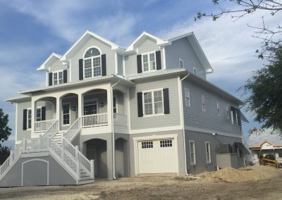 Large two-story light gray house with white trim, storm shutters, front porch, stairs, and double garage in Morehead City, North Carolina under a blue sky.