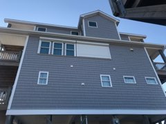 ATLANTIC BREEZE STORM SHUTTERS ROLLING SHUTTERS44 Gray three-story house on stilts with multiple windows and a durable white roll shutter, perfect for North Carolina coastal weather.