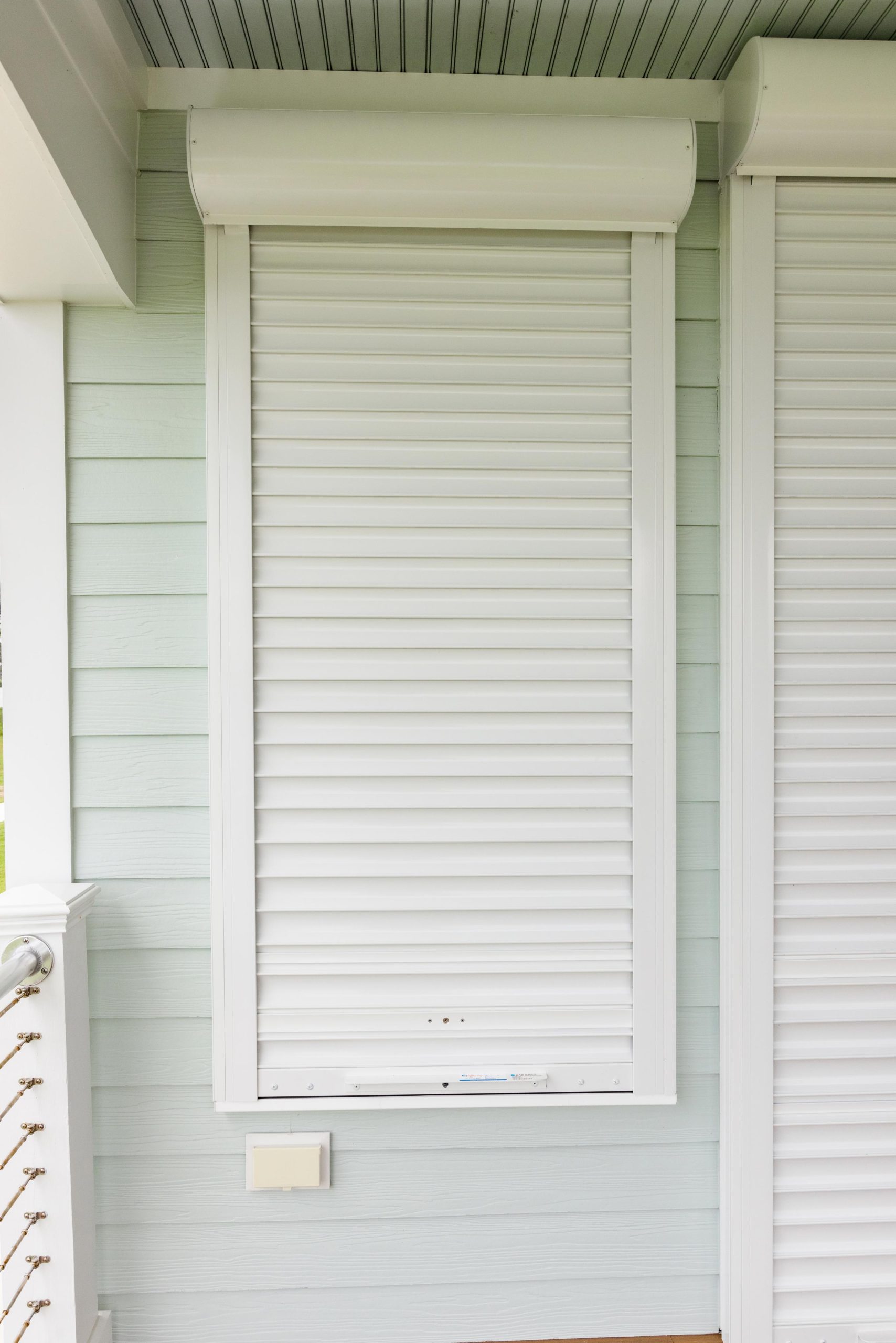 Atlantic-Breeze-Storm-Shutters-9458 Atlantic Breeze Storm Shutters: White roll-down hurricane shutter closed over a window on a light blue house exterior.