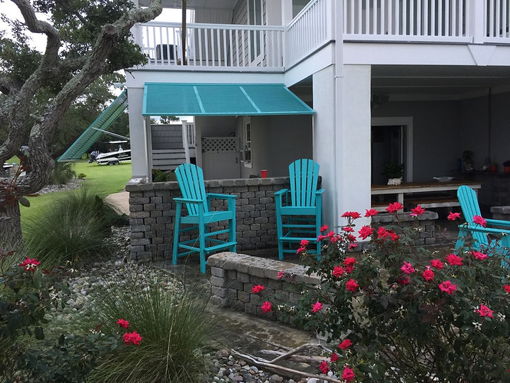 ebbd78_0a3a6c6d711d4666bb08c443b Two turquoise patio chairs and a table rest on a stone terrace near blooming red flowers and a white house, offering storm protection for relaxing days in Morehead City, North Carolina.