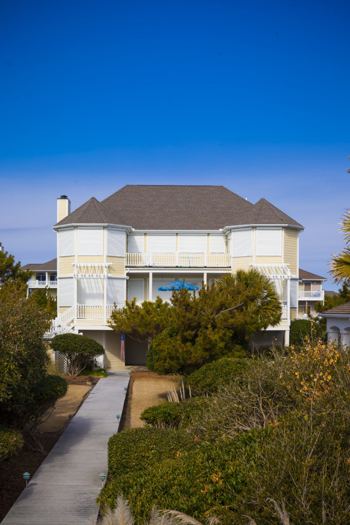 A large, two-story beach house with balconies and roll shutters for storm protection, surrounded by bushes and a clear blue sky above.