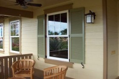 Light yellow house exterior with a window, green shutters, wooden chairs, and a wall-mounted light fixture.