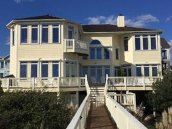 ebbd78_2c9ff0a589b6495590c6a6033 Large yellow beach house with many windows and roll shutters, featuring a wooden walkway leading up to the front deck in Morehead City, North Carolina.