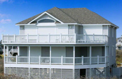 Two-story white house with wraparound porch and railings, set against a blue sky.