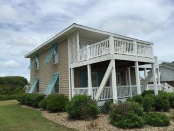 Two-story house with blue shutters, white trim, and wraparound balconies, surrounded by bushes and grass—featuring Storm Shutters for added storm protection against Atlantic Breeze winds.