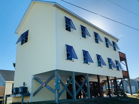 ebbd78_b77e1f4e959242abaa36cdc66 Yellow house on stilts with blue window shutters, elevated to protect from flooding under a clear sky—classic Atlantic Breeze style in Morehead City, North Carolina.