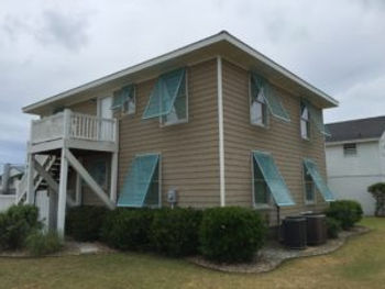 ebbd78_cd0085d78bfb4cabaa20e5030 Two-story beige house with teal roll shutters for storm protection, lush shrubs, and an exterior staircase on a cloudy day.