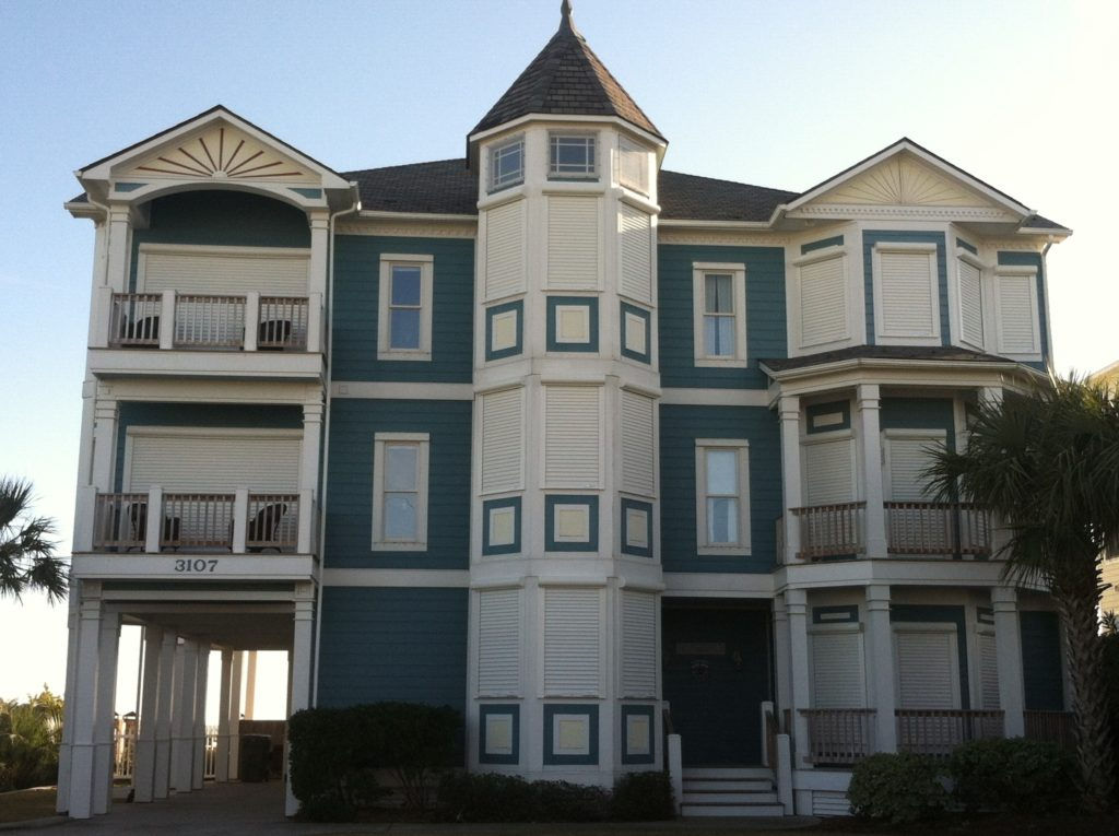 ABSS Large blue and white three-story beach house with balconies, turret, and palm trees in front.
