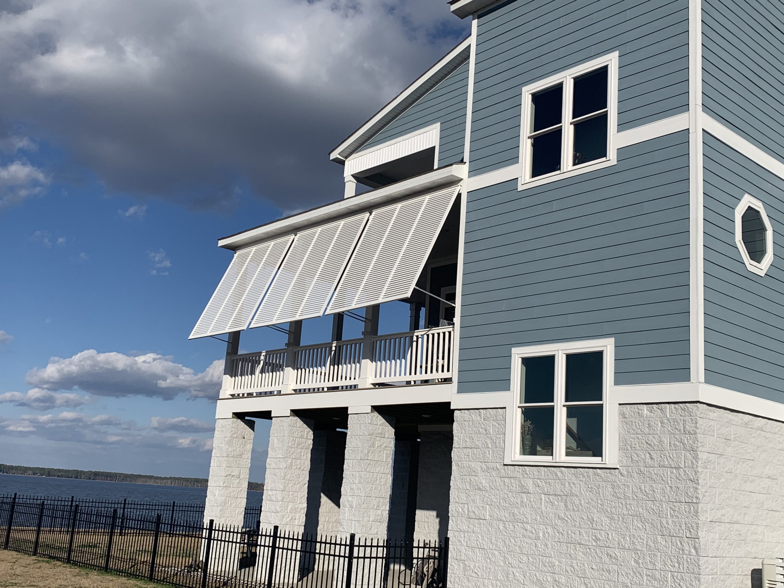 Blue house with white awnings covering a balcony, next to a fence and overlooking a body of water.