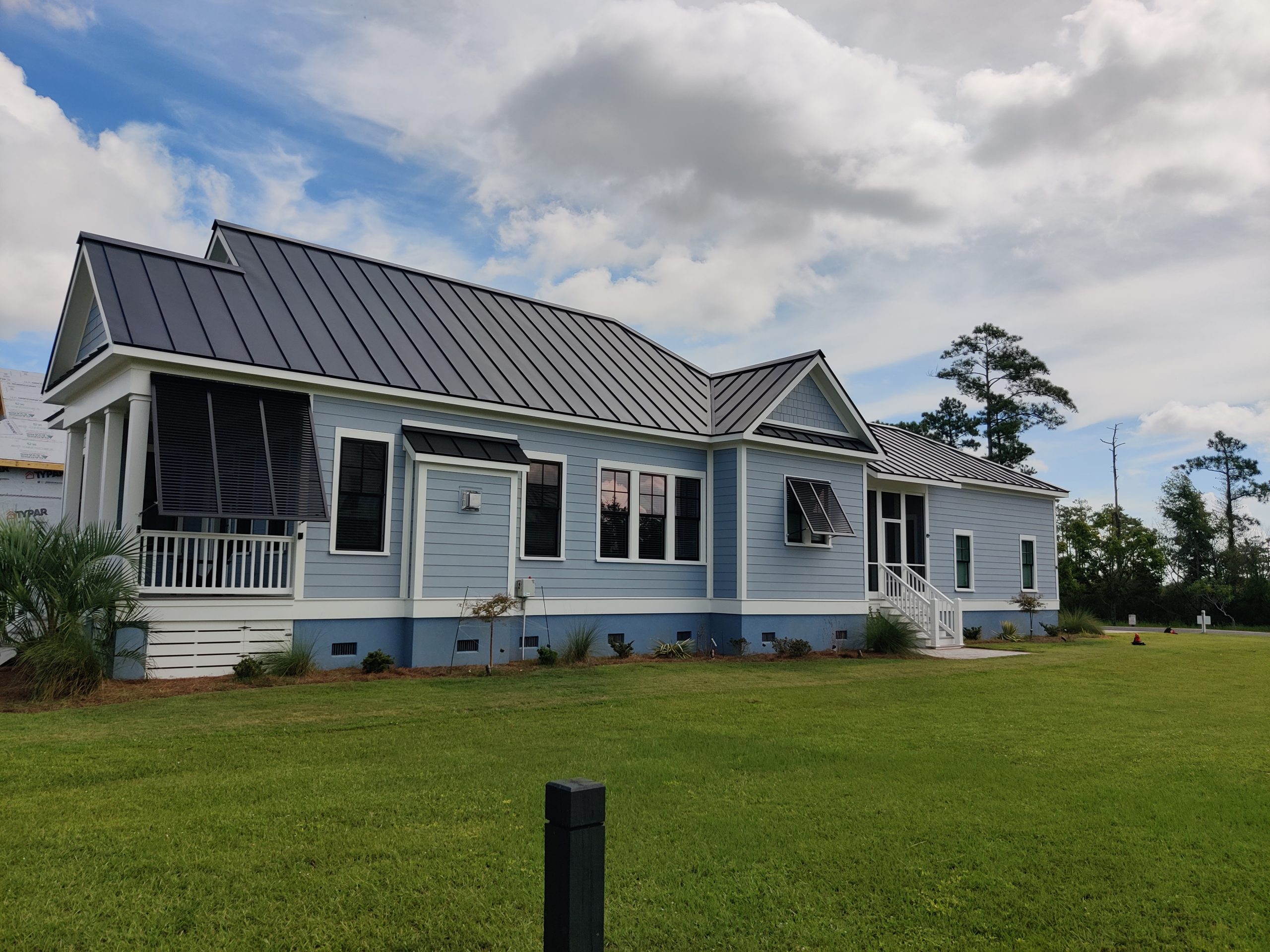 Light blue house with black metal roof, white trim, shutters, and a large green lawn under a partly cloudy sky.