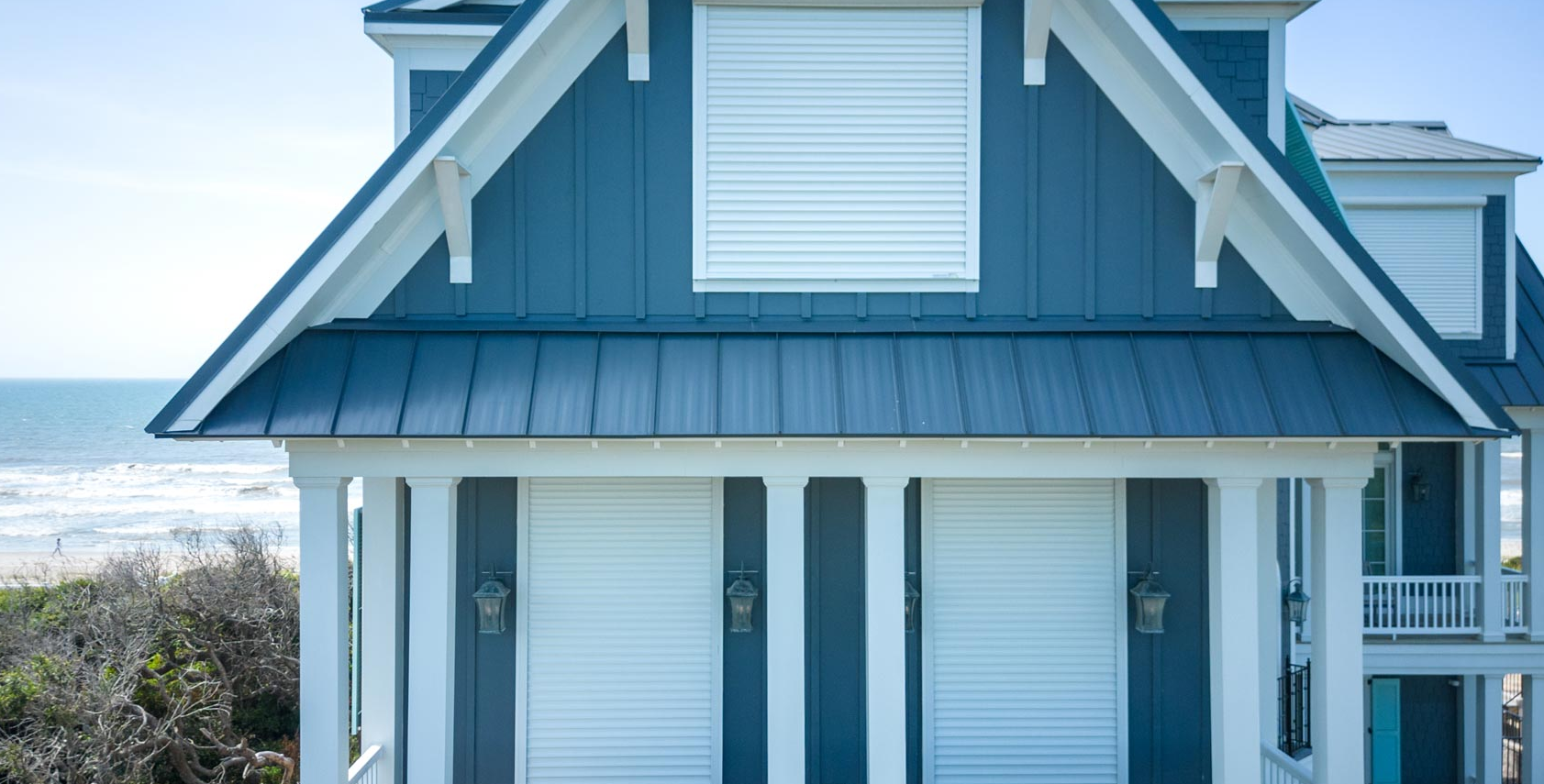 A blue beach house with shutters closed, overlooks the ocean on a clear day.