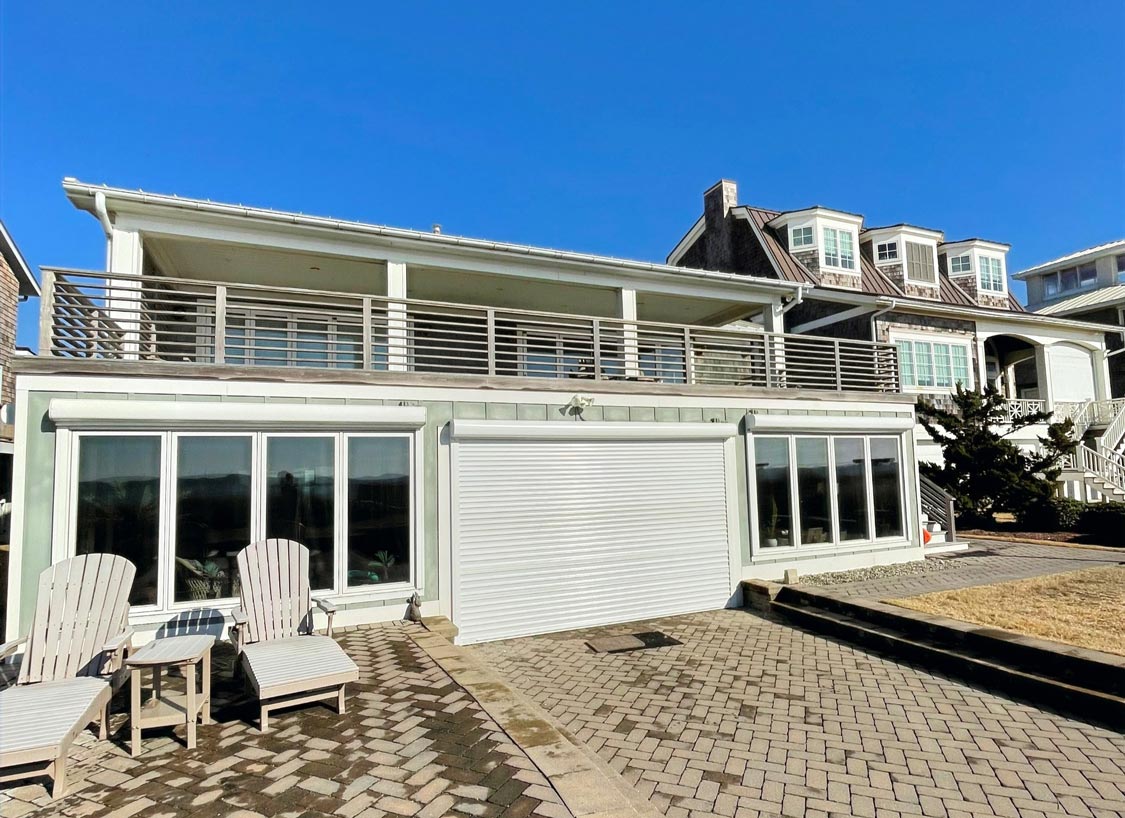 Modern beach house with large windows, upper balcony, and patio seating on a sunny day.